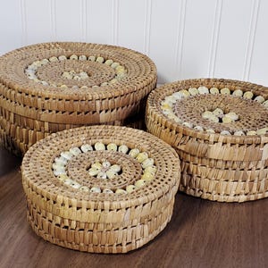 Puede incluir: Tres cestas redondas tejidas de diferentes tamaños. Las cestas están hechas de paja de color natural y presentan un patrón espiral decorativo en las tapas, acentuado con pequeñas cuentas de color claro. Una manzana verde está a la izquierda de las cestas.