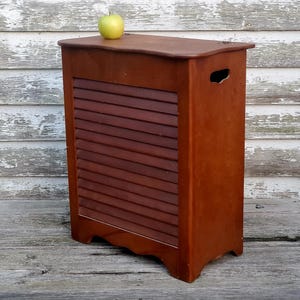 May include: A wooden cabinet with a slatted front and a curved base. The cabinet is stained a warm brown color and has a handle cut out on the side. A green apple sits on top of the cabinet.