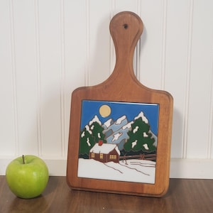 May include: Wooden cutting board with a handle, featuring a ceramic tile insert. The tile depicts a winter scene with a cabin, snow-covered mountains, and a full moon. The board is made of light brown wood.
