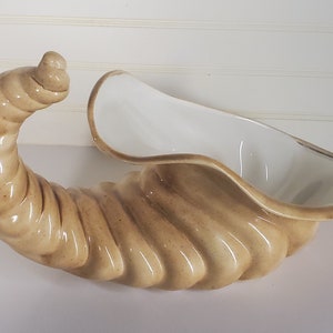May include: A ceramic cornucopia in a light brown colour with a white interior. The cornucopia is shaped like a curled horn with a textured surface.