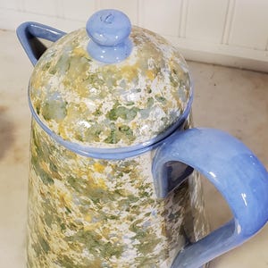 以下が含まれることがあります： 緑、黄色、白の斑点模様の仕上げが施された陶器製のピッチャー。このピッチャーには、無地の水色のハンドル、つまみ付きの水色の蓋、そして容器の上部開口部を囲む水色の縁取りが特徴として備わっています。