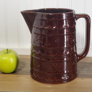 May include: A dark brown ceramic pitcher with a handle and spout, decorated with embossed floral and wave patterns. A green apple sits to the left of the pitcher. The pitcher is likely used for serving beverages.