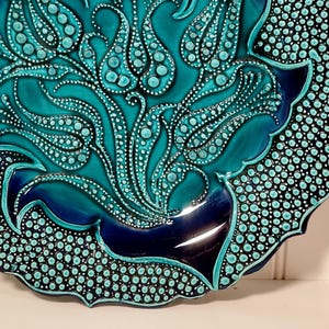 May include: A decorative ceramic plate with a turquoise and navy blue floral design. The plate features a scalloped edge and a glossy finish. The floral pattern is raised and detailed with small white dots.