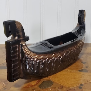 Könnte beinhalten: Dekorative, bootsförmige Vase aus Keramik in Braun- und Schwarztönen. Die Vase hat eine glänzende Oberfläche und weist detaillierte Reliefmuster an den Seiten auf. Die Enden des Bootes sind erhöht und haben ein stilisiertes Design.