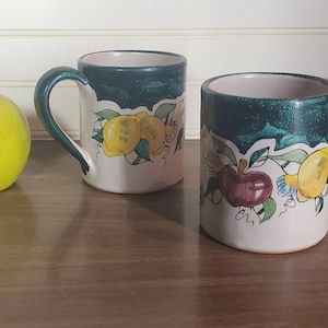 May include: Two ceramic mugs with green and white designs featuring lemons, apples, and leaves. The mugs have a handle on the right side.