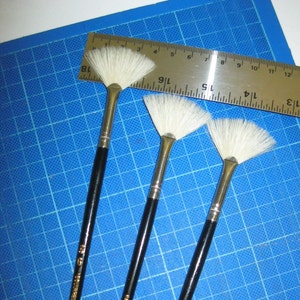 Może przedstawiać: Trzy czarne, wachlarzowe pędzle z białymi włosiem. Pędzle są ułożone w kształt trójkąta na niebieskiej powierzchni w kratkę. Na uchwytach nadrukowany jest tekst "SQUIRREL CO. CHINA".