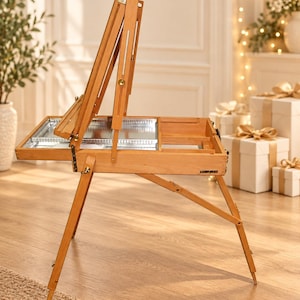 May include: A light brown wooden tabletop easel with an adjustable frame and a built-in storage compartment featuring a metal interior tray. The easel includes folding legs and metal hardware, designed to hold a canvas upright for painting.