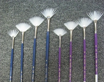 May include: A collection of fan-shaped paint brushes with white bristles and long handles. The handles are either blue or purple, with silver ferrules. The brush tips are white. The brushes are arranged in a row.