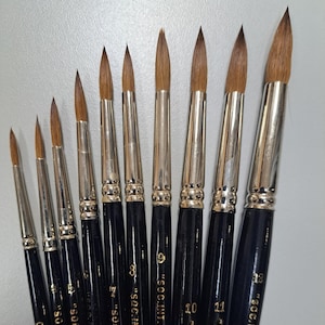 Peut inclure: Un ensemble de dix pinceaux ronds avec des manches noirs et des inscriptions dorées. Les pinceaux sont numérotés de 0 à 12 et portent l'inscription "SOC.INT.CAN.MTL.". Les pinceaux sont disposés par ordre de taille, du plus petit au plus grand.