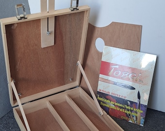 Könnte beinhalten: Ein Holzkoffer für ein Künstler-Staffelei mit einem Scharnierdeckel und einem Griff. Der Koffer hat drei Fächer im Inneren und ist geöffnet, um die Fächer zu zeigen. Neben dem Koffer befindet sich eine Packung Einweg-Malpaletten.