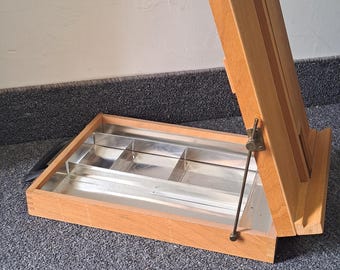 Könnte beinhalten: Ein Holzstaffelei mit einem Metalltablett für Farben. Die Staffelei ist geöffnet und das Tablett ist sichtbar. Die Staffelei ist aus hellem Holz und hat ein schlichtes Design.