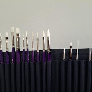 Może przedstawiać: Zestaw pędzli malarskich z białym włosiem i fioletowymi trzonkami, starannie ułożonych w czarnym organizerze z tkaniny. Pędzle różnią się rozmiarem i kształtem, w tym okrągłymi, płaskimi i skośnymi końcówkami, odpowiednimi do różnych technik malarskich.