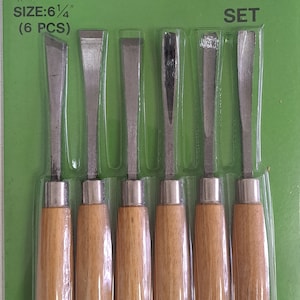 Può includere: Un set di sei strumenti per intagliare con manici in legno. Gli strumenti sono confezionati in una confezione blister di plastica trasparente su uno sfondo verde. La confezione riporta la scritta "CARVING TOOL SET" e "SIZE: 6 1/4" (6 PCS)".
