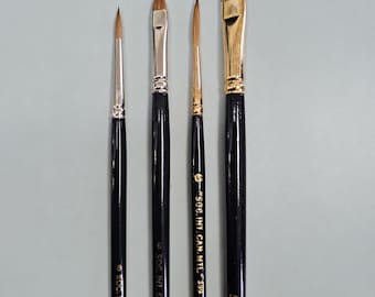 May include: A set of four artist paint brushes with glossy black wooden handles and gold-toned metal ferrules. The brushes feature varying tip shapes, including round and flat styles, with gold text printed on the handles reading SOC. INT. CAN. MTE.