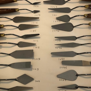 Puede incluir: Un juego de 22 cuchillos de paleta de pintura de metal con mangos de madera. Los cuchillos están dispuestos en dos columnas, con los números 1001 a 1108 escritos junto a cada cuchillo.