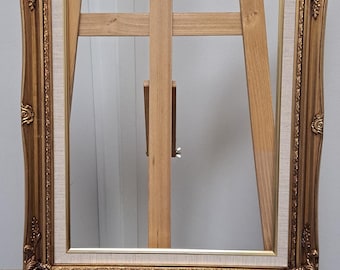 May include: A gold-colored, ornate picture frame with a cream-colored inner border, displayed on a wooden artist's easel. The frame has detailed carvings and a rectangular shape. The easel is made of light-colored wood.