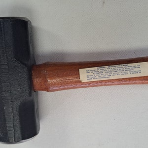 May include: A black sledgehammer with a wooden handle. The handle has a warning label that reads "WARNING: WEAR SAFETY GLASSES".