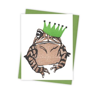 May include: A brown toad wearing a green crown with black accents. The toad is sitting on a white background.