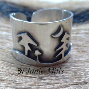 May include: A silver ring with a design of two trees and a crescent moon. The ring is on a light brown furry surface. By Jamie Mills.