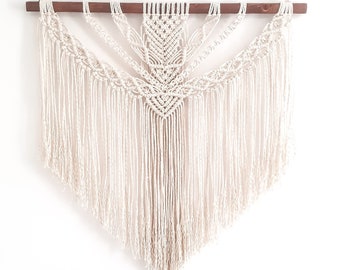 Puede incluir: Un tapiz de macramé blanco con un diseño geométrico y flecos. El tapiz está sujeto a una varilla de madera.