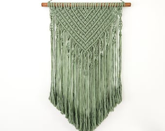 May include: Sage green macrame wall hanging with a geometric design. The wall decor features a wooden dowel and long, flowing fringe. This handmade piece adds a bohemian touch to any room.