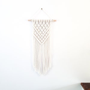 May include: White macrame wall hanging with a geometric pattern and fringe. The hanging is attached to a wooden dowel.