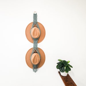 May include: A macrame hat rack with two brown hats hanging on it. The rack is made of light blue cord and has a fringe at the bottom. The rack is mounted on a white wall. A small green plant is in a white pot on a wooden shelf to the right of the rack.