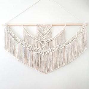 Puede incluir: Un tapiz de macramé blanco con un patrón geométrico y flecos. El tapiz está sujeto a una varilla de madera.