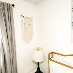 May include: A cream-colored macrame wall hanging with a geometric design is mounted on a white wall. A black lamp with a white lampshade sits on a white nightstand. A gray curtain hangs to the left.