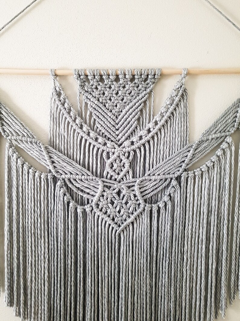 May include: A gray macrame wall hanging with a geometric pattern and fringe. The hanging is attached to a wooden dowel.