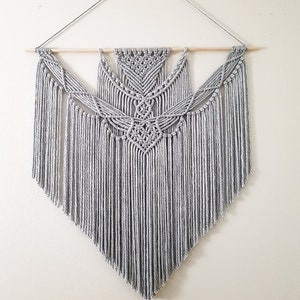 May include: A gray macrame wall hanging with a geometric pattern and fringe. The hanging is attached to a wooden dowel rod.