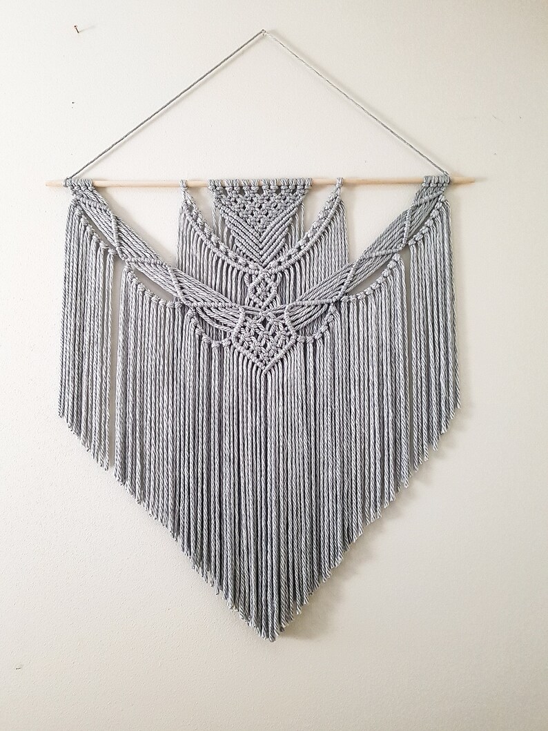 May include: A gray macrame wall hanging with a geometric pattern and fringe. The hanging is attached to a wooden dowel rod.