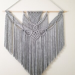 May include: A gray macrame wall hanging with a geometric pattern and fringe. The hanging is made with thick cotton cord and is attached to a wooden dowel.