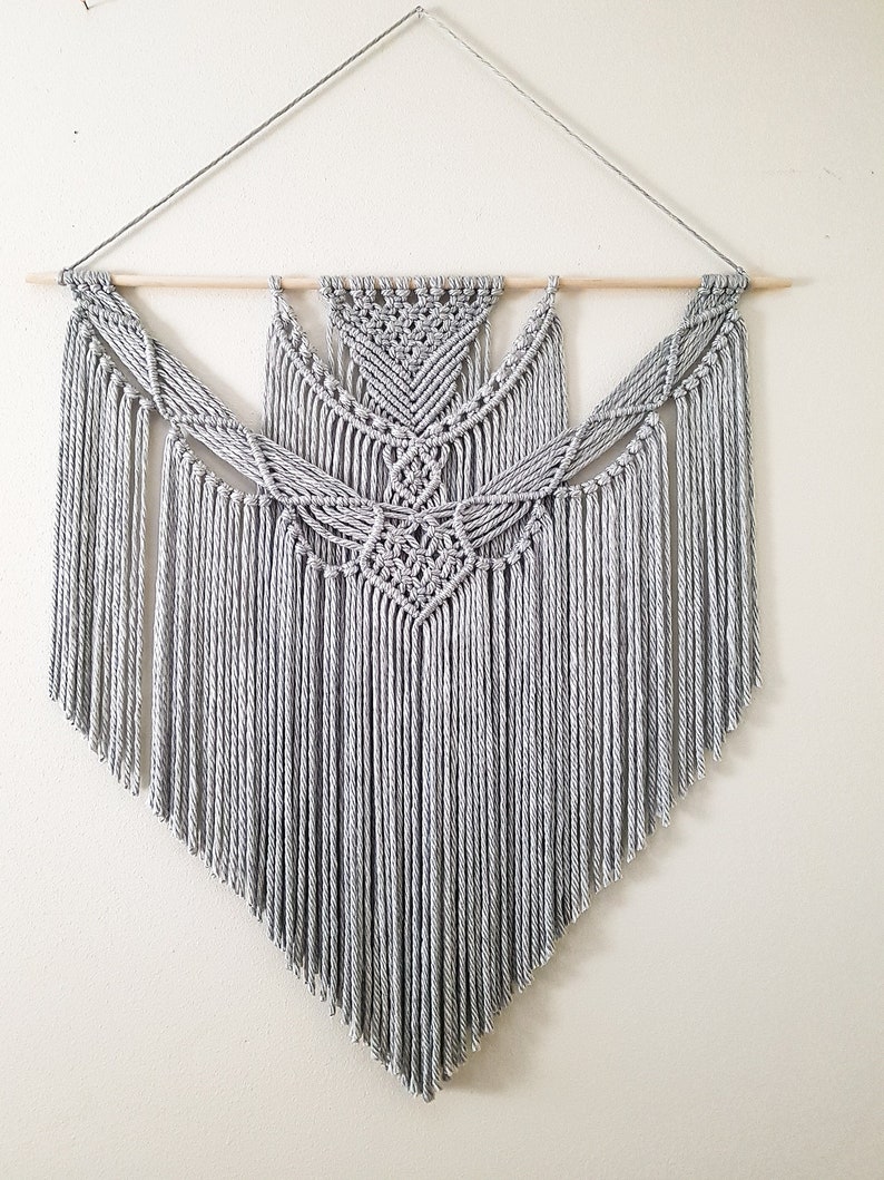 May include: A gray macrame wall hanging with a geometric pattern and fringe. The hanging is made with thick cotton cord and is attached to a wooden dowel.