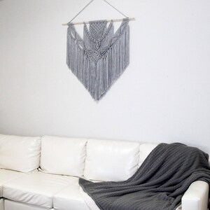 May include: A white couch with a gray throw blanket draped over it. A gray macrame wall hanging with a wooden dowel is hanging on the wall behind the couch.