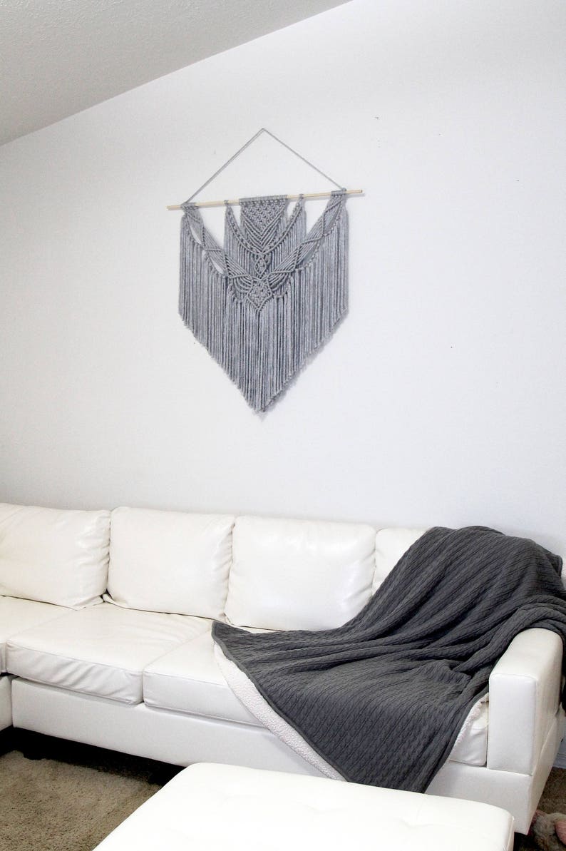 May include: A white couch with a gray throw blanket draped over it. A gray macrame wall hanging with a wooden dowel is hanging on the wall behind the couch.