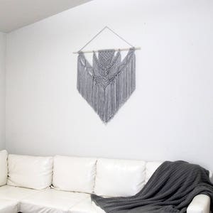 May include: A gray macrame wall hanging with a geometric design hangs on a white wall above a white leather sectional sofa with a gray throw blanket.