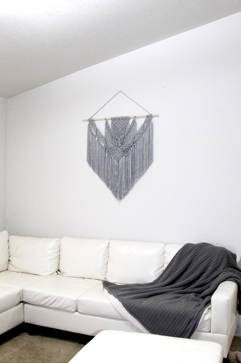 May include: A gray macrame wall hanging with a geometric design hangs on a white wall above a white leather sectional sofa with a gray throw blanket.