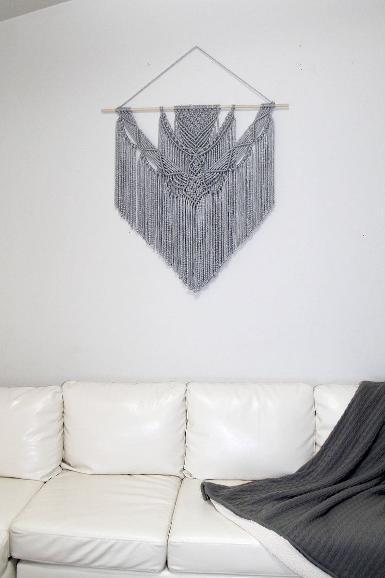 May include: A gray macrame wall hanging with a geometric design. The hanging is made with thick yarn and has a wooden dowel at the top.