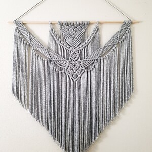 May include: A gray macrame wall hanging with a geometric design. The hanging is made with thick cotton cord and has a wooden dowel at the top.