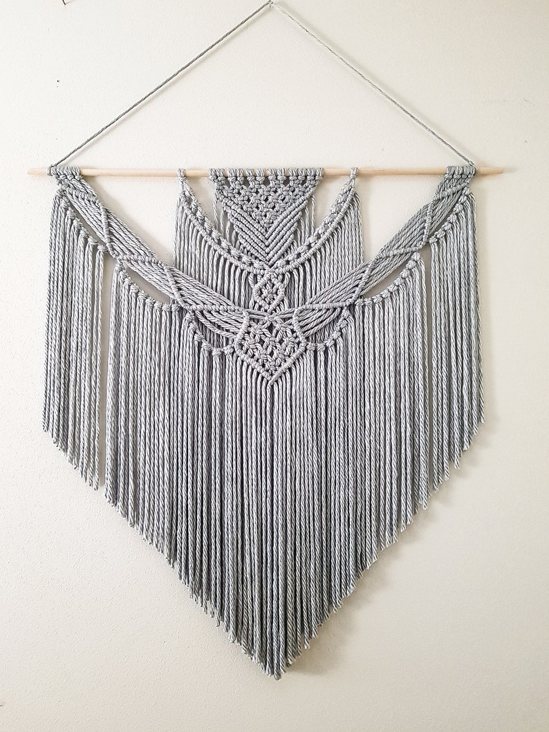 May include: A gray macrame wall hanging with a geometric design. The hanging is made with thick cotton cord and has a wooden dowel at the top.
