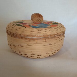 May include: A round woven wicker basket with a wooden knob lid. The lid is painted with a colorful floral design.