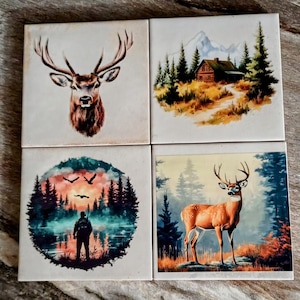 May include: Four square ceramic tiles with nature-themed illustrations. One tile features a deer head, another a cabin in the mountains, a third a hunter by a lake, and the last a deer in a forest. The tiles are white with colorful images.