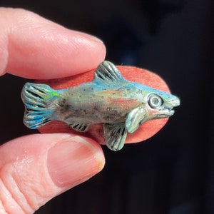 May include: A small, detailed, hand-painted ceramic sculpture of a trout fish on a wooden base. The fish is painted in shades of blue, green, and brown with iridescent accents.