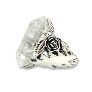 May include: A clear quartz crystal with a silver-colored metal accent. The metal piece is shaped like a stylized rose with leaves. The crystal is a light, translucent color, and the metal has intricate details.