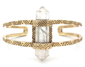 May include: A gold-colored cuff bracelet featuring a clear quartz crystal. The bracelet has an open design with geometric patterns and a central setting that holds the crystal. The crystal is a clear, elongated shape.