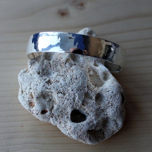 May include: A silver hammered bangle bracelet with a smooth, polished finish. The bracelet is displayed on a white rock.
