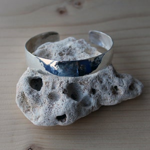 May include: A silver hammered bangle bracelet, resting on a white rock.