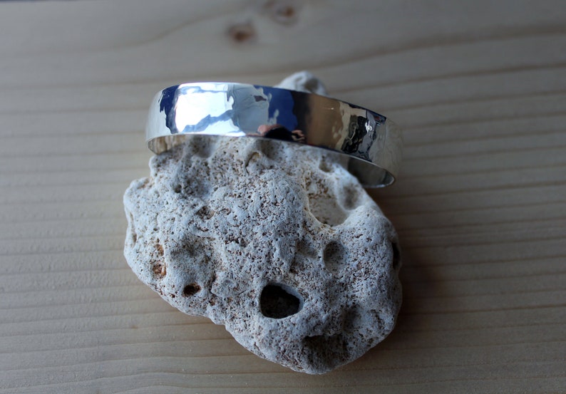 May include: A silver hammered bangle bracelet with a smooth, polished finish. The bracelet is displayed on a white rock.
