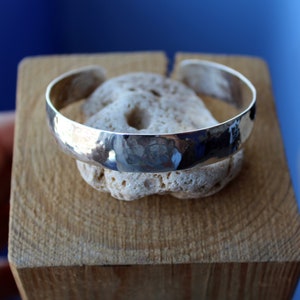 May include: A silver hammered cuff bracelet with a smooth, rounded surface. The bracelet is displayed on a piece of light brown wood.